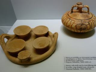 A gauche, petites tasses pour les offrandes rituelles sur un plateau, à côté, un vase en forme d'anneau et en arrière plan, un pot à étrier. Ces pièces proviennent de Gournes Pediada (1300-1200 av. JC.).